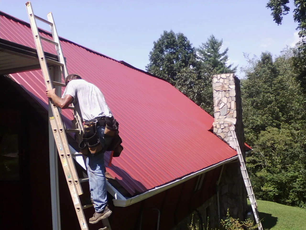 Roofer on a ladder beside a red metal roof and stone chimney — Soffit & Fascia Repair by Roofing Brothers in Toppenish